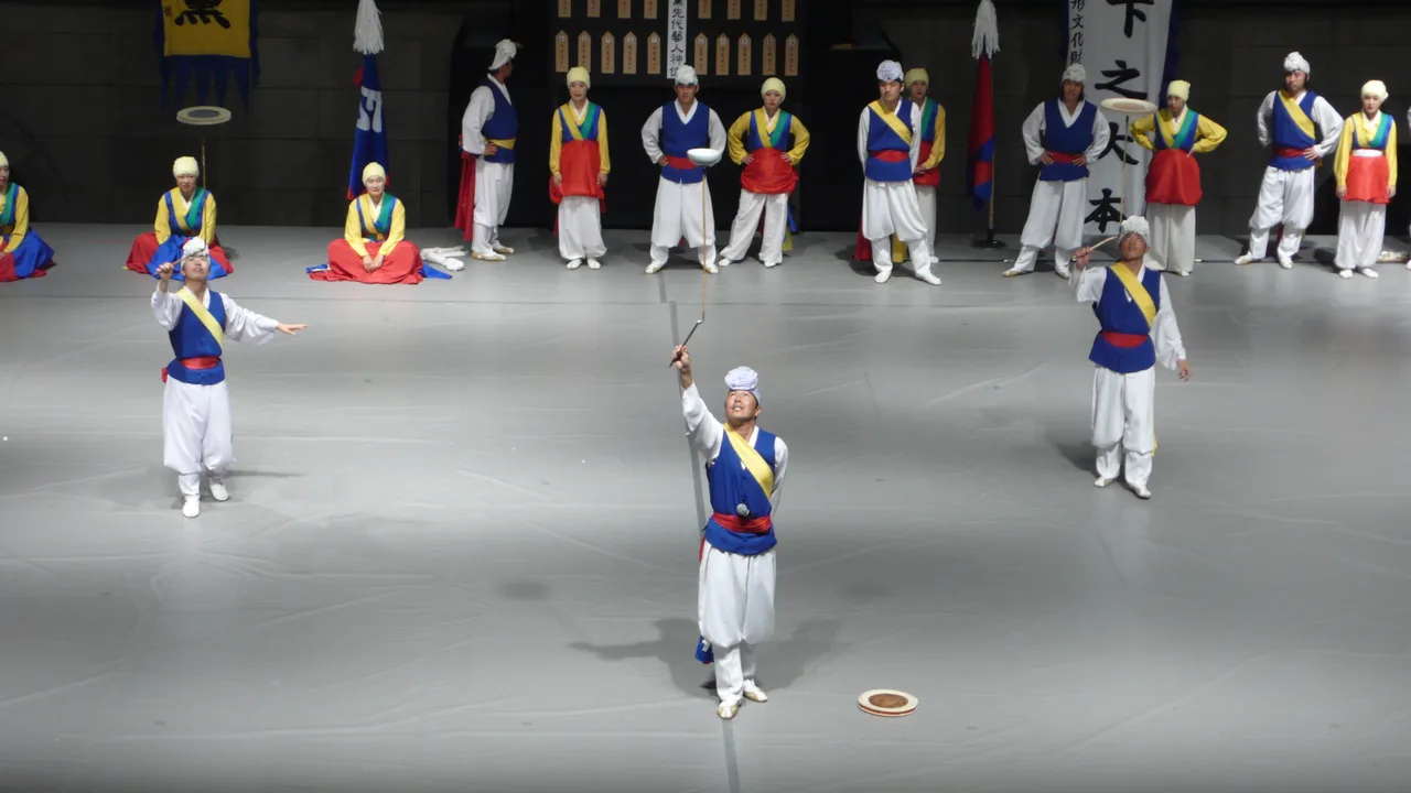 막대 위에 접시를 올려 돌리는 버나잽이 공연자와 뒤에서 이를 지켜보는 전통 복장의 공연단