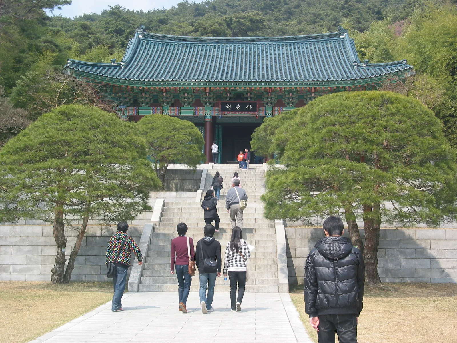 소나무 사이 돌계단 위에 현충사라고 쓰인 현판이 걸린 정문 건물이 보이고 관람객들이 계단을 오르는 모습
