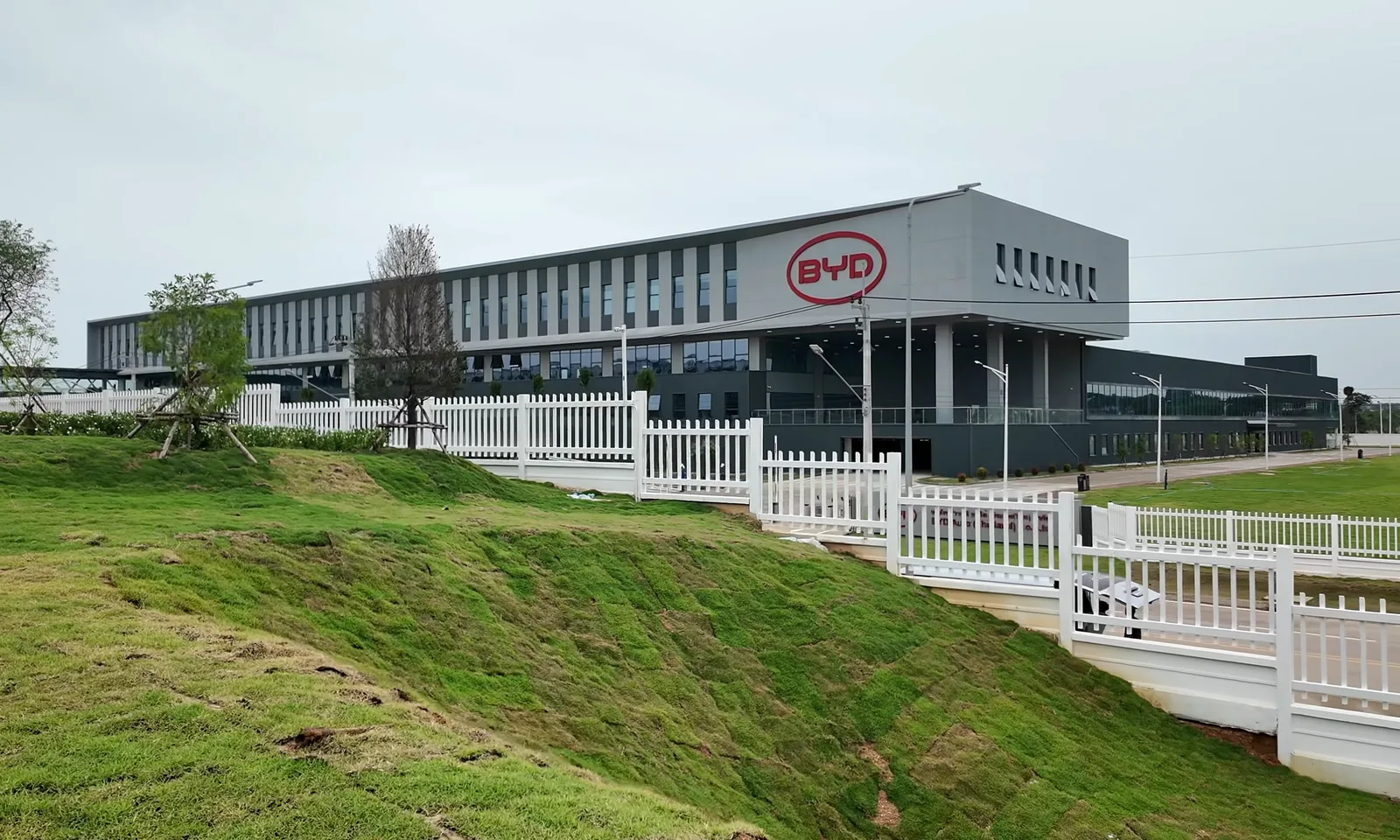 흐린 하늘 아래 BYD 로고가 크게 걸린 회색 톤의 자동차 공장 건물, 앞쪽에 잔디 언덕과 흰색 울타리가 늘어서 있는 모습
