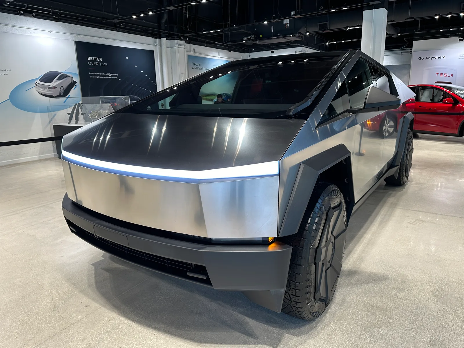 테슬라 매장 내부에 은색 사이버트럭이 전시돼 있고, 뒤쪽으로 빨간색 테슬라 차량과 TESLA 로고가 보이는 모습