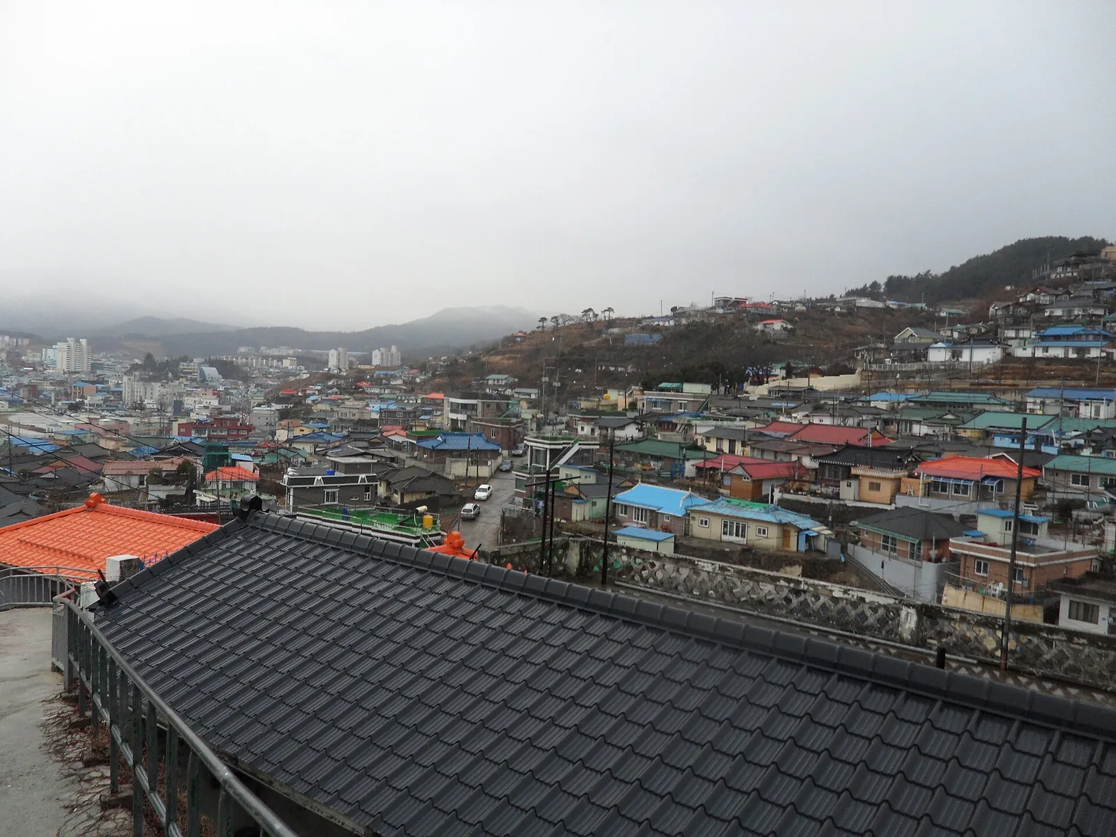 흐린 하늘 아래 언덕을 따라 낮은 지붕의 집들이 빼곡하게 들어선 마을 전경, 앞쪽에 기와지붕이 보인다