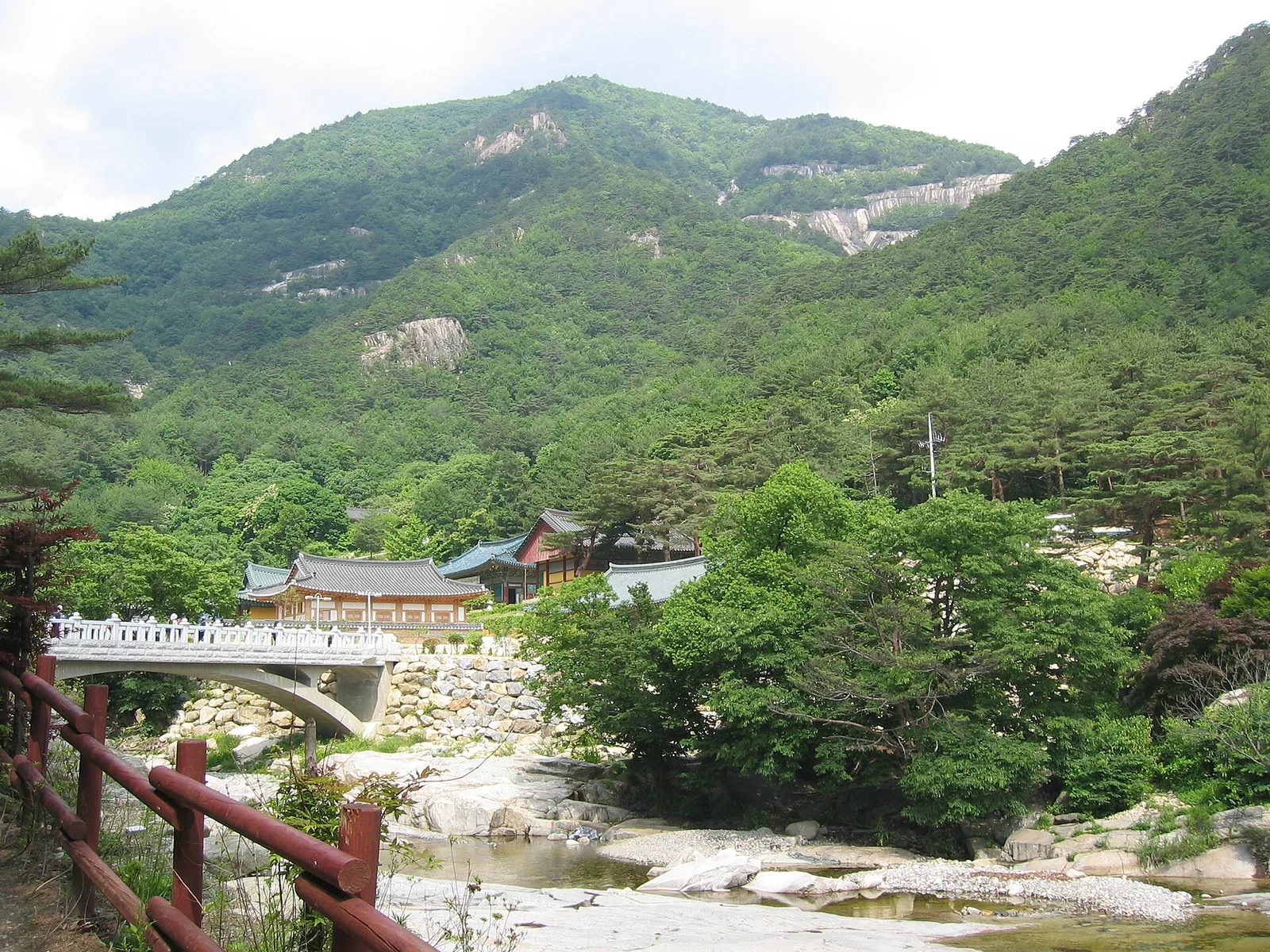 푸른 산자락 아래 계곡을 가로지르는 다리와 그 너머 기와지붕의 사찰 건물들이 보이는 풍경