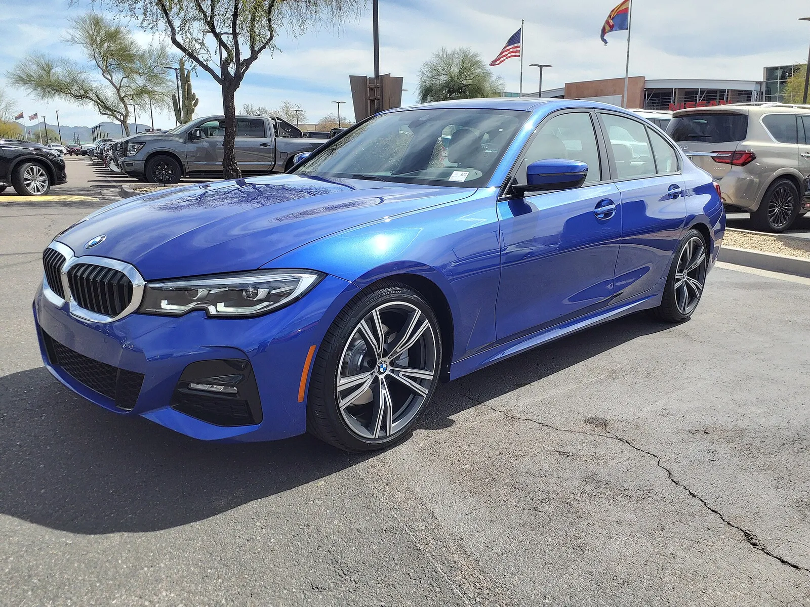 파란색 BMW G20 세대 3시리즈 330i의 전면과 측면, 주차장 배경에 성조기가 걸려 있다