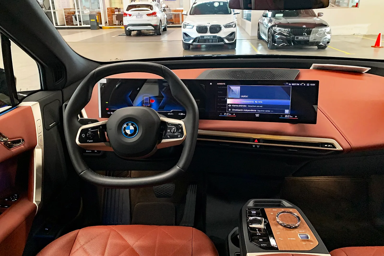 BMW iX 실내 운전석, 곡면 디스플레이와 육각형 스티어링 휠, 주황색 가죽 대시보드가 보인다