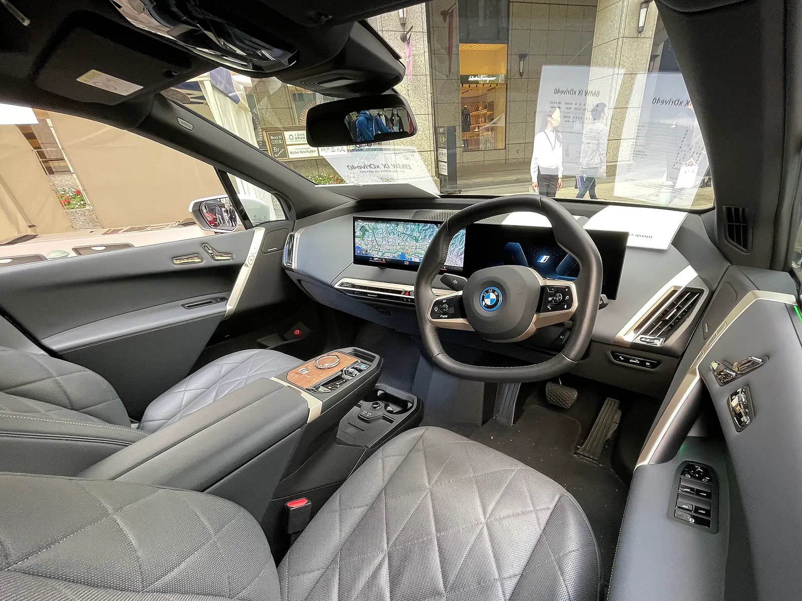 우측 핸들 BMW iX xDrive40 실내, 육각형 스티어링 휠과 곡면 디스플레이, 회색 퀼팅 시트가 보인다