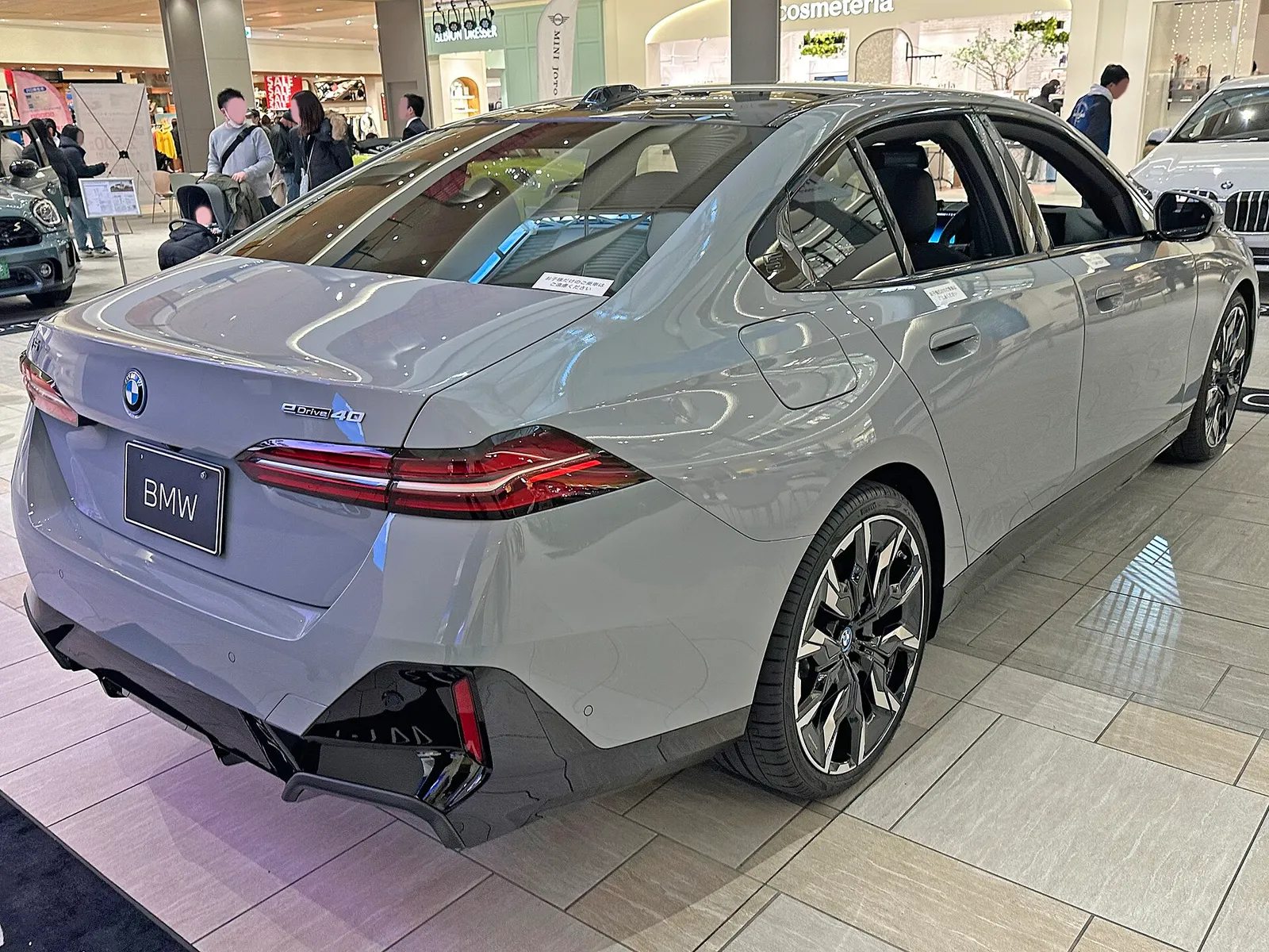 실내 전시장에 놓인 회색 BMW i5 eDrive40 M 스포츠 후면 3/4, 낮게 이어진 테일램프와 대형 멀티스포크 휠이 보인다