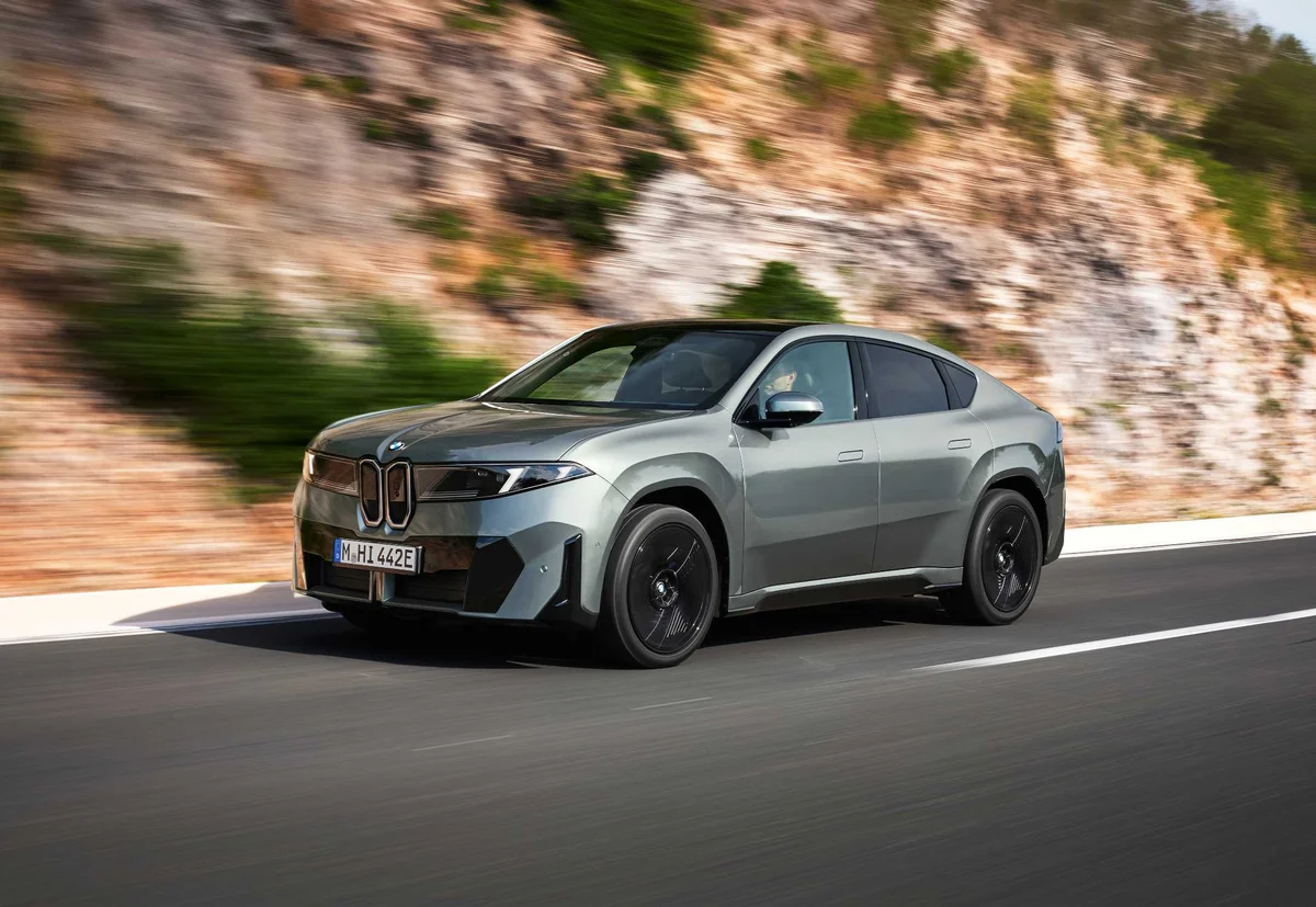 회색 BMW iX4 50 xDrive가 암석 절벽 옆 도로를 달리는 전면 3/4 모습