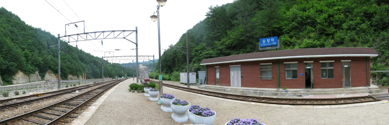 산으로 둘러싸인 승부역 승강장과 붉은 벽돌 역사, 보라색 꽃 화분이 놓인 모습