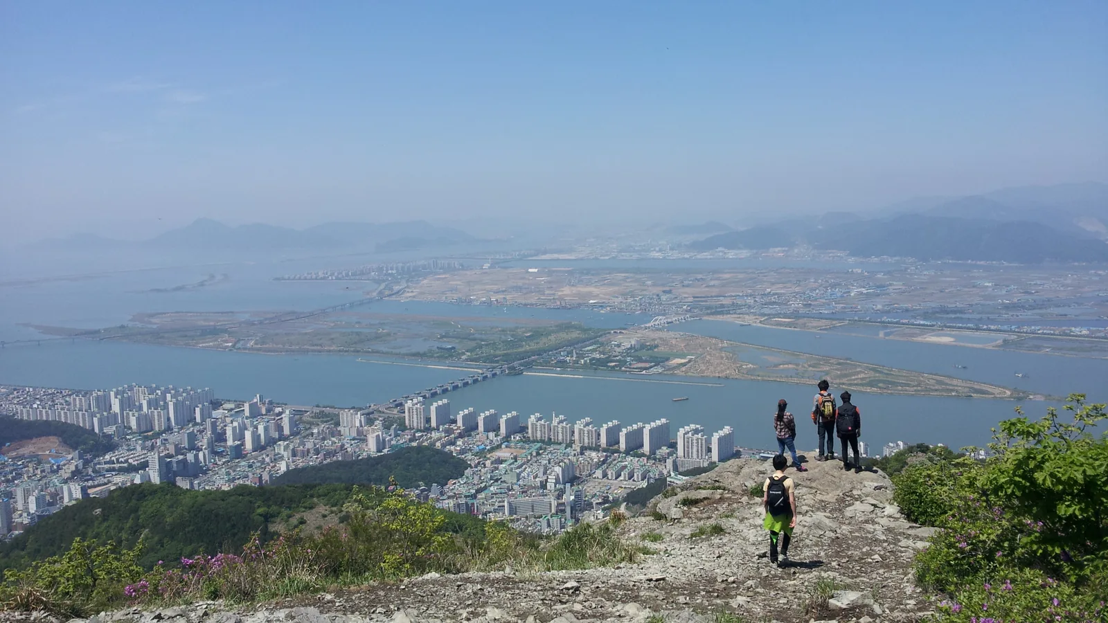 산 정상에서 내려다본 낙동강 하구 전경, 강이 여러 갈래로 갈라지며 바다와 만나고 그 사이로 삼각주 습지와 다리, 도심이 펼쳐져 있다