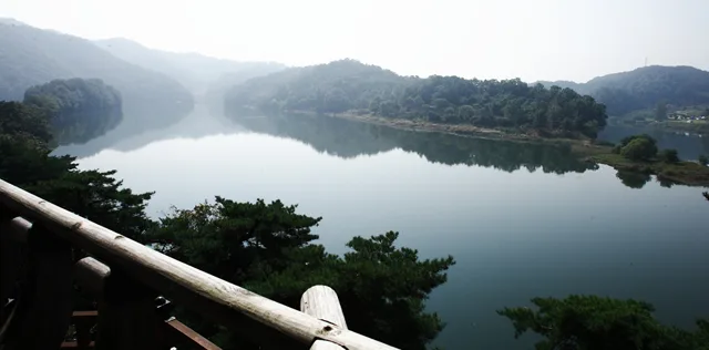 통나무 난간 너머로 소나무 사이로 잔잔한 호수와 물굽이를 따라 이어진 숲이 내려다보이는 풍경