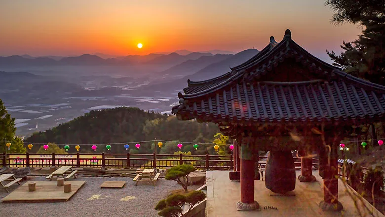 주황빛 새벽하늘 아래 겹겹이 이어진 산줄기 위로 해가 떠오르고, 오른쪽에 전통 기와지붕의 종각이 서 있는 풍경