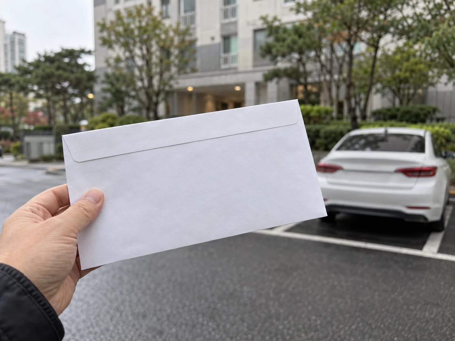 아파트 주차장 앞에서 한 손에 글자가 없는 흰 봉투를 들고 있고 뒤에 흰색 승용차가 주차돼 있는 모습
