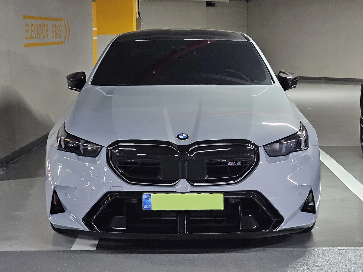 주차장에 서 있는 회색 BMW M5 G90 세단의 앞모습, 큰 키드니 그릴과 M 엠블럼이 보인다