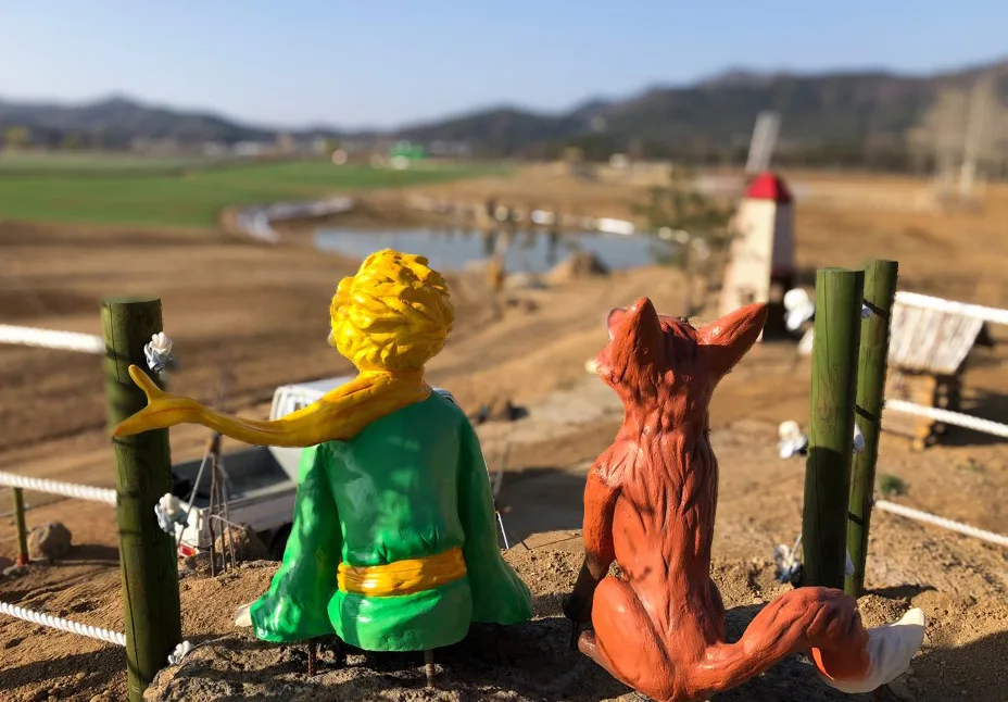 꽃밭 한가운데 세워진 어린왕자와 여우 조형물, 노을을 바라보고 있다