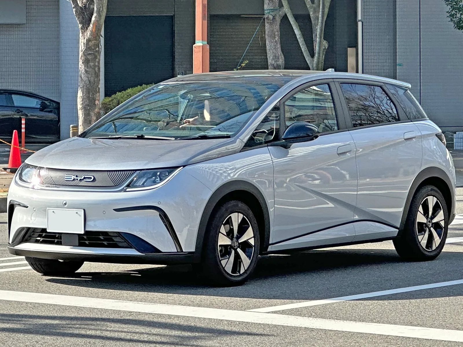 일본 도심 도로에 주차된 은색 BYD 돌핀의 전면 및 측면 모습, BYD 레터링이 새겨진 그릴과 번호판이 보인다