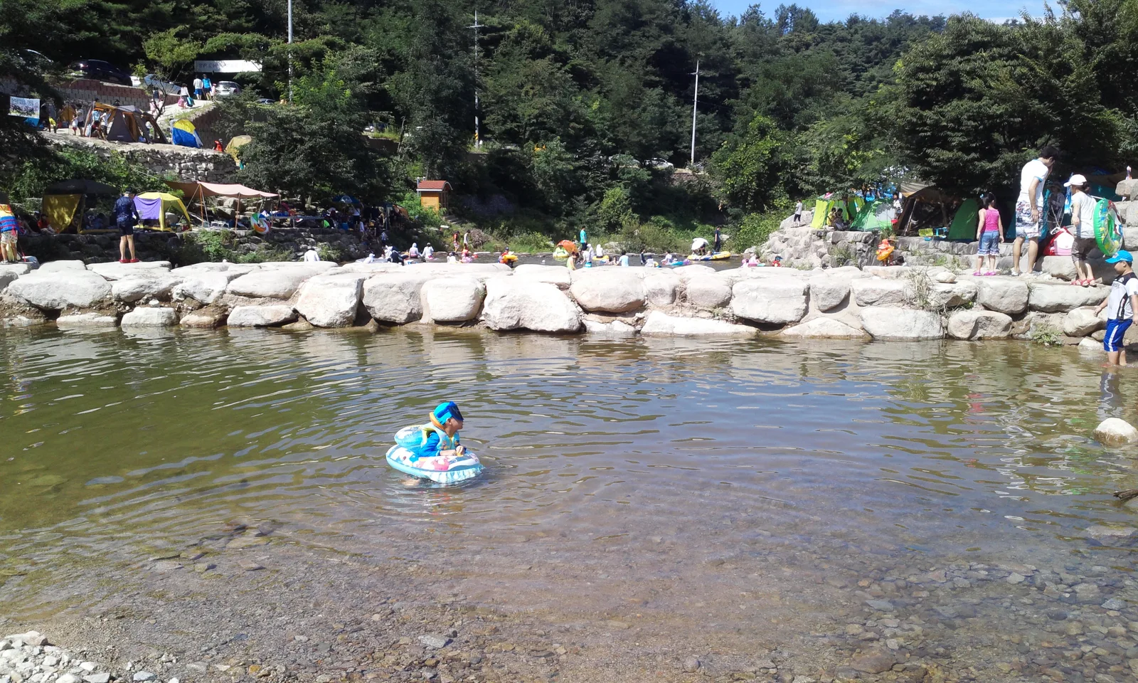 팔공산 자락 계곡에서 튜브를 타고 물놀이를 즐기는 아이와 주변 야영객들, 돌로 쌓은 둔치와 울창한 숲이 배경