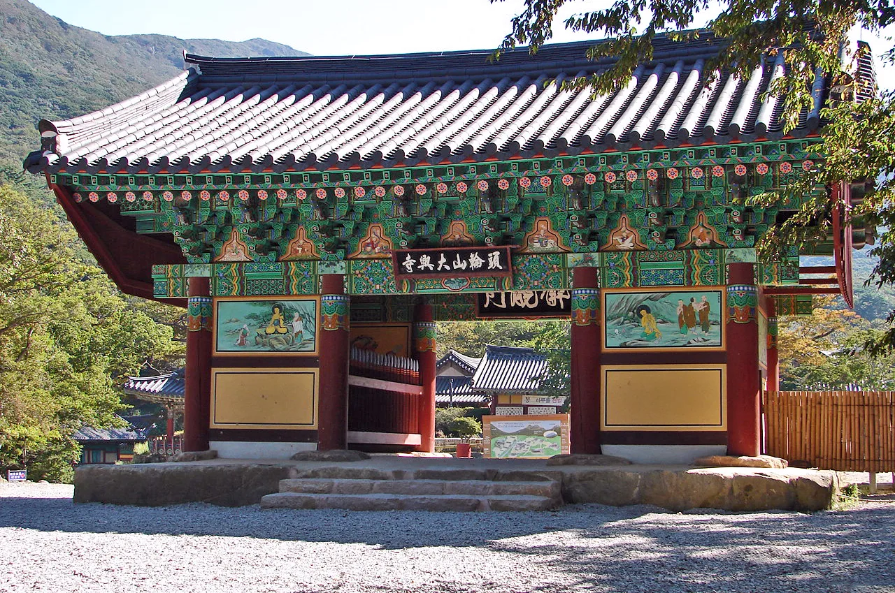 화려한 단청을 입힌 사찰 전각이 자갈 마당 위에 서 있고 주변으로 숲이 우거져 있다