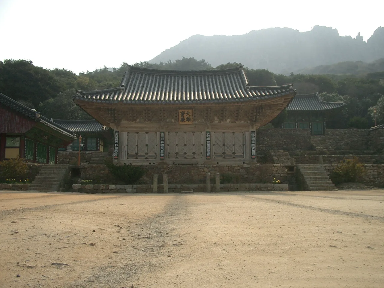 넓은 흙 마당 너머로 단청 없는 목조 법당이 있고 그 뒤로 뾰족한 바위 능선이 옅은 안개 속에 이어진다
