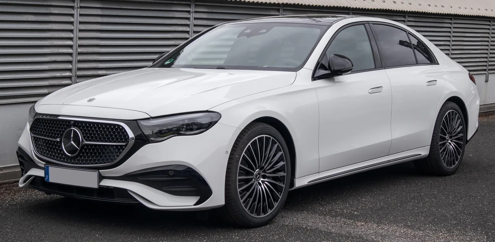 실외에 주차된 흰색 메르세데스-벤츠 E-Class E200 AMG 라인의 정측면 모습, 세로형 그릴과 AMG 전용 휠이 보인다