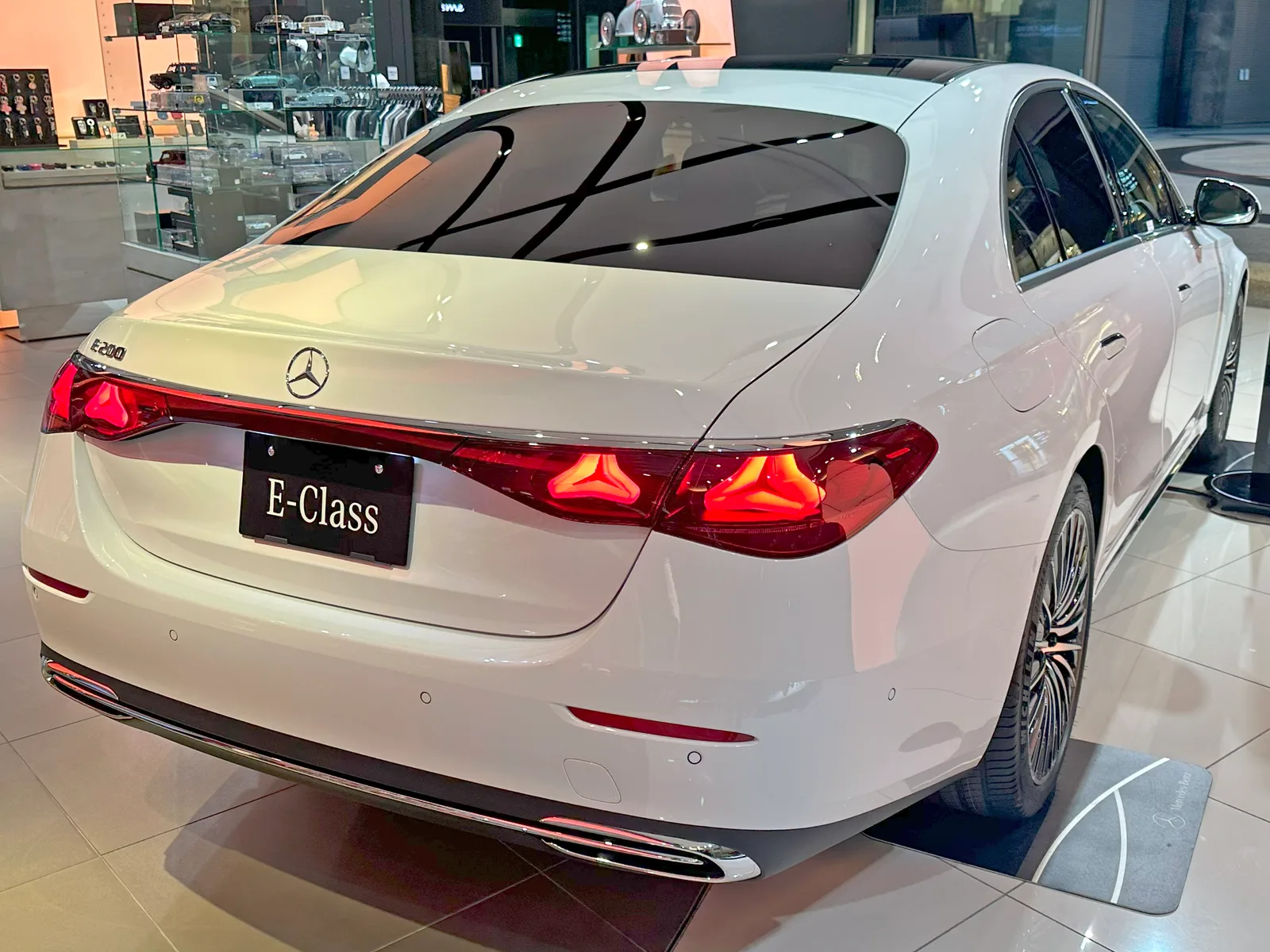 전시장에 놓인 흰색 메르세데스-벤츠 E-Class E200의 후면, 트렁크에 E-200 배지와 E-Class 번호판 프레임이 보이는 모습