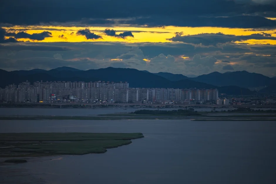 구름 사이로 노을빛이 남은 하늘 아래 낙동강 하구 너머로 도시와 산 능선이 보이는 저녁 풍경
