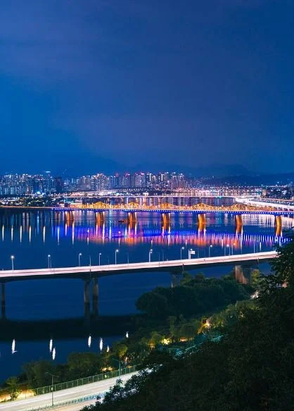 강 위 다리의 조명과 도시 야경이 보이는 세로 구도의 사진