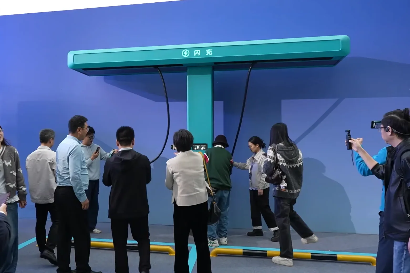 파란색 배경의 전시장에서 사람들이 스마트폰으로 촬영하며 지켜보는 가운데, 청록색 T자형 BYD 플래시 충전 시연 장비가 설치돼 있는 모습