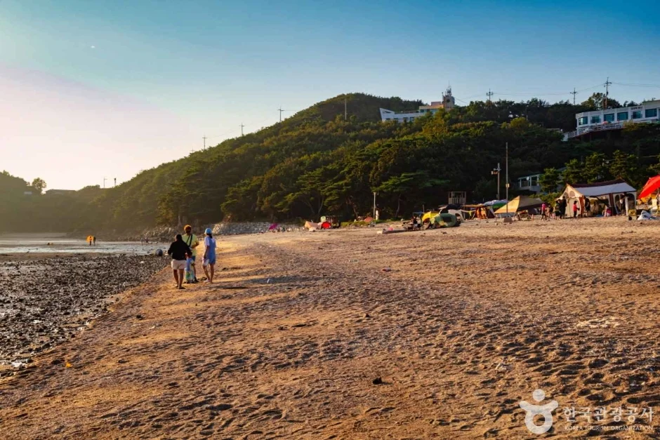 낮게 비치는 햇빛 아래 산자락을 낀 모래 해변을 걷는 사람들
