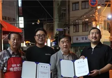 정장을 입은 남성 두 명이 김해뒷고기푸드페스타 명예홍보대사 위촉장을 들고 다른 두 명과 나란히 서 있는 모습