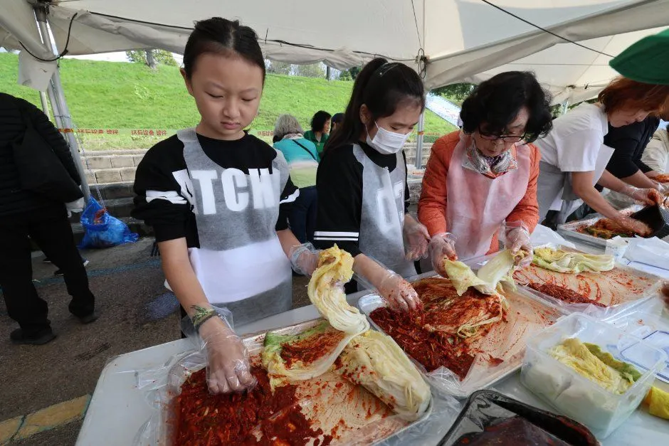 천막 아래에서 어린이와 가족이 배추에 양념을 버무려 김치를 담그는 체험 모습