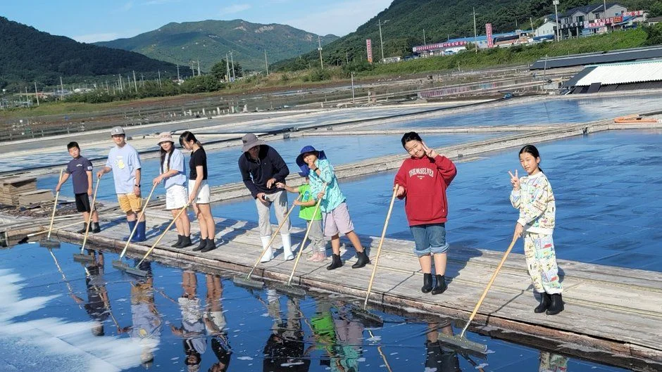 염전 위 나무 통로에서 아이들이 고무래로 소금을 모으는 체험 장면
