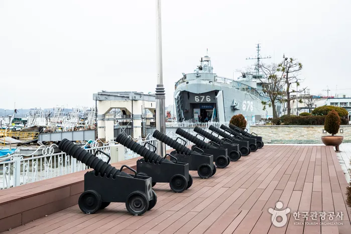 바다 쪽 나무 데크에 여러 문의 대포가 나란히 놓이고 뒤로 함선이 보이는 진포해양테마공원