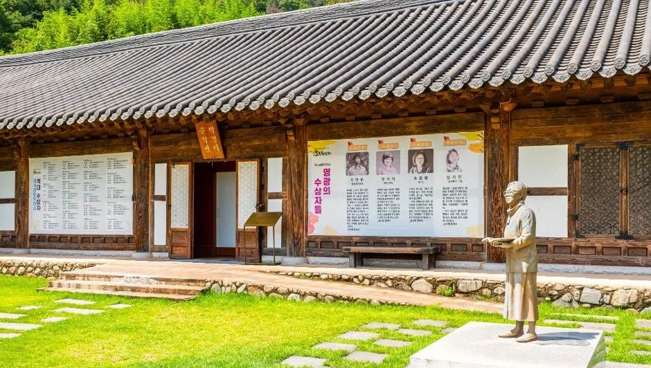 기다란 한옥 앞에 인물 동상과 안내판이 서 있는 모습