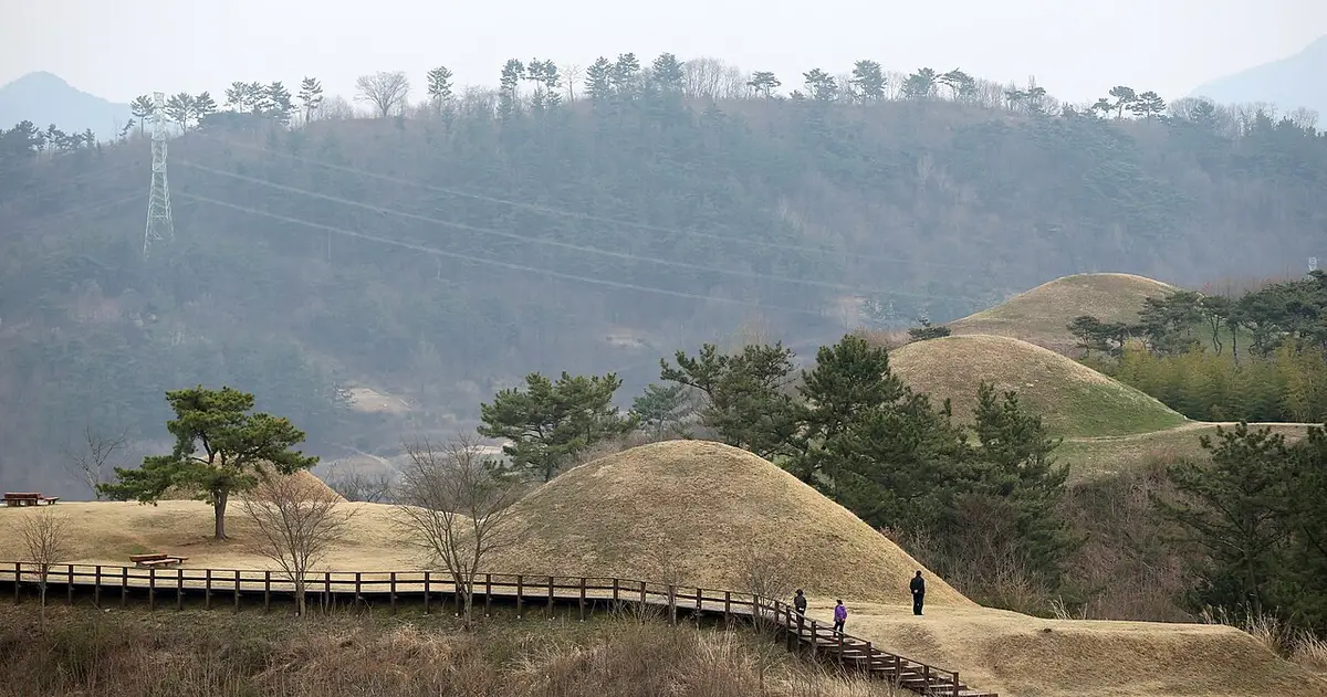 누런 잔디의 고분과 앙상한 나무, 목재 탐방로를 걷는 사람들