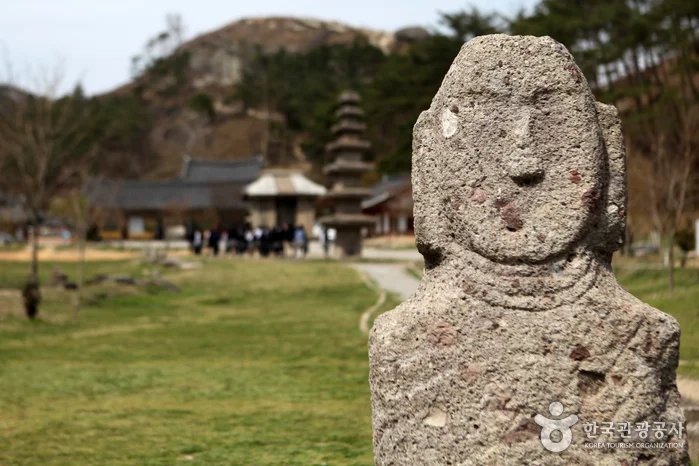 거친 표면의 석불 얼굴 클로즈업과 뒤로 보이는 탑과 사찰 건물