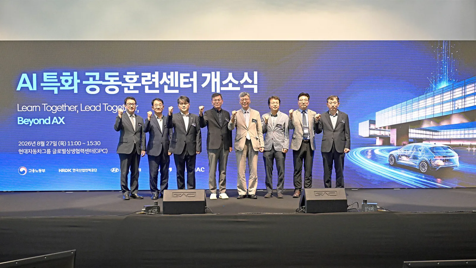 AI 특화 공동훈련센터 개소식 무대 클로즈업, 관계자 8명이 주먹을 쥐고 포즈를 취한 모습, 뒤로 'Learn Together, Lead Together, Beyond AX' 슬로건과 개소식 일시·장소 문구가 크게 표시된 스크린