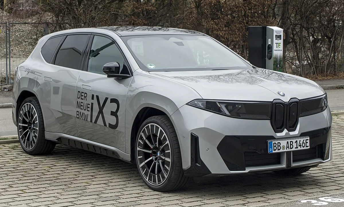 독일 전시장 앞 보도에 선 은색 BMW iX3의 전측면, 차체 옆에 'DER NEUE BMW iX3' 문구가 붙어 있다