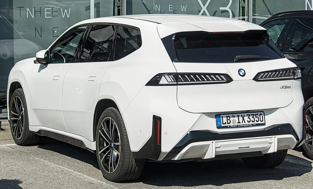 독일 전시장 앞에 선 흰색 BMW iX3의 후측면, 가로로 길게 이어진 테일램프가 보인다