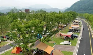 넓은 잔디밭 캠핑장에 텐트와 캐노피가 늘어서 있고 뒤로 산이 보이는 전경