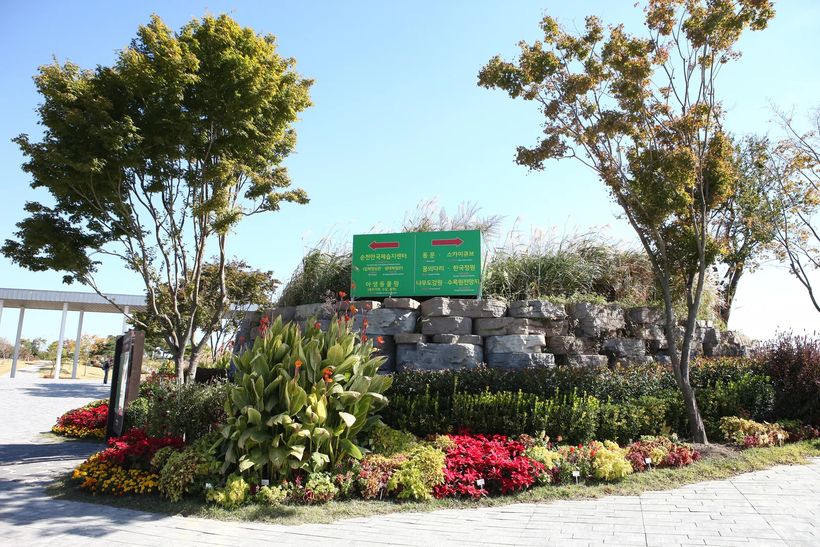 화단과 억새 사이로 방향을 안내하는 초록색 안내판이 세워진 정원 입구, 뒤로 단풍 든 나무가 보인다
