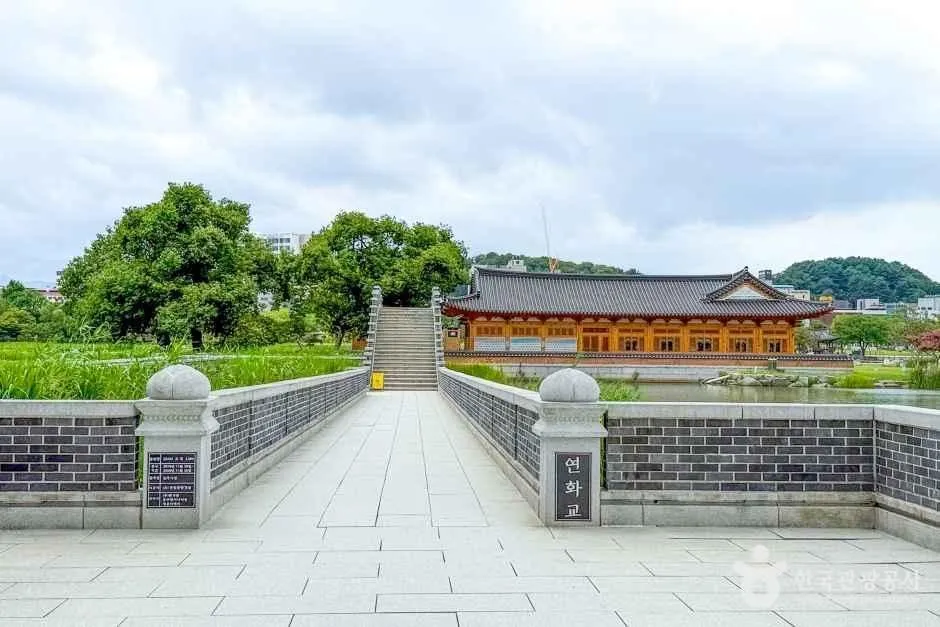 돌난간이 있는 넓은 보행 다리 끝에 기와지붕의 한옥 건물이 보이고 양옆으로 수생식물이 우거진 덕진공원 풍경