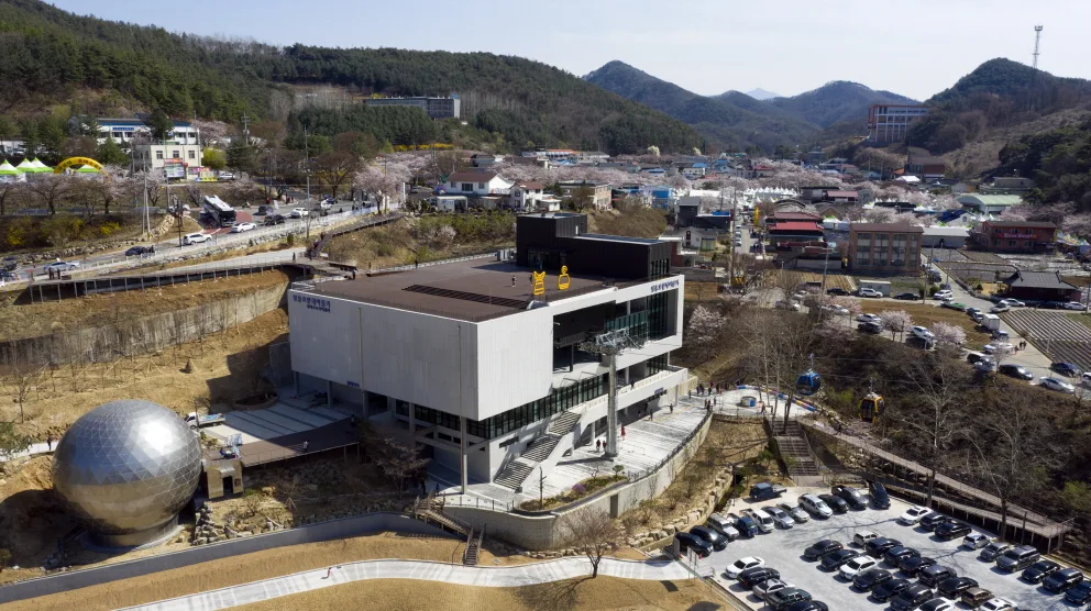 둥근 시네마360 조형물과 넓은 주차장을 갖춘 청풍호반케이블카 물태리역(하부역) 건물, 산 위로 향하는 캐빈들이 보인다