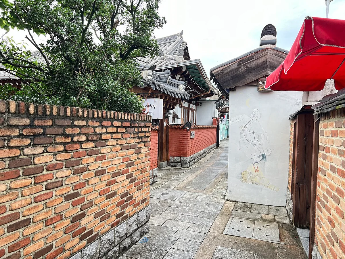 붉은 벽돌 담 옆으로 돌바닥 골목이 이어지고 한옥 지붕이 보이는 모습