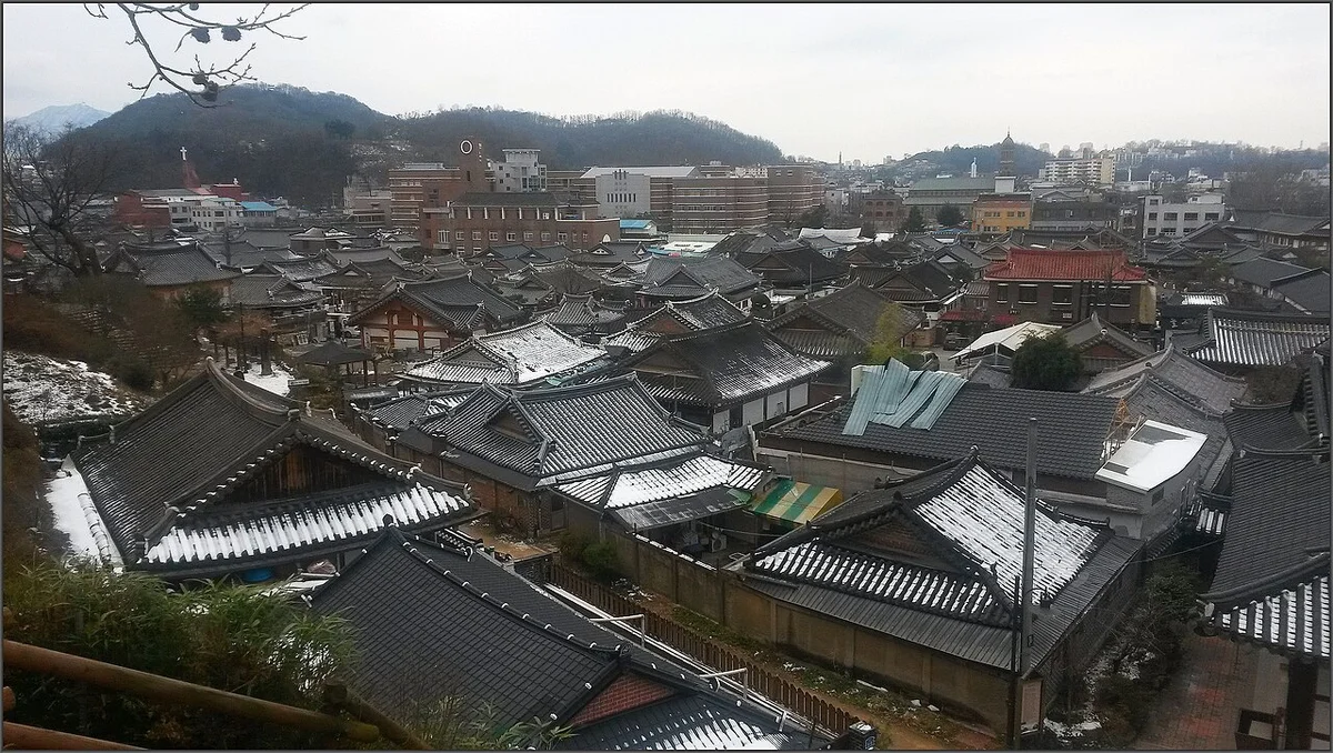 기와지붕 위에 눈이 쌓이고 멀리 산과 시가지가 보이는 한옥마을 전경