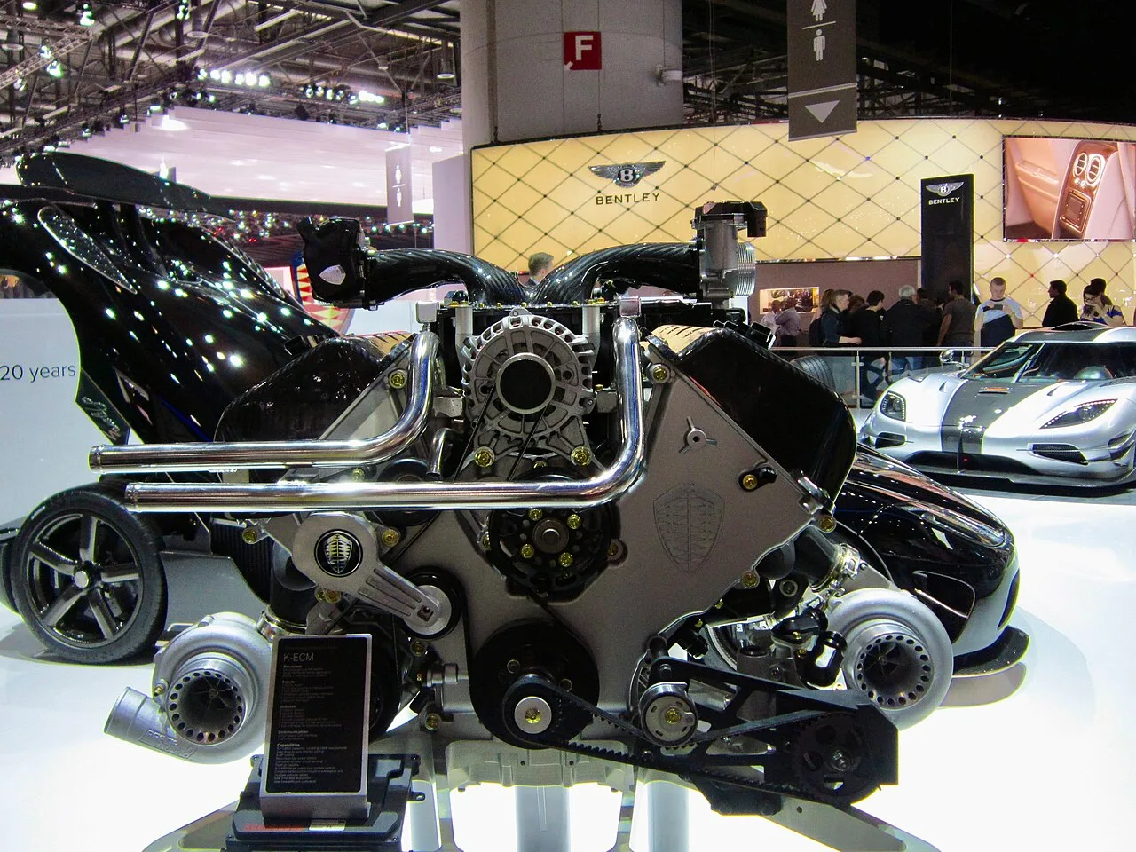 제네바 모터쇼 전시대에 놓인 쾨닉세그 원:1의 5.0L 트윈터보 V8 엔진을 근접 촬영한 사진, 좌우로 트윈터보차저와 배기 매니폴드가 보이고 뒤편에 실버 색상 쾨닉세그 차량이 보인다