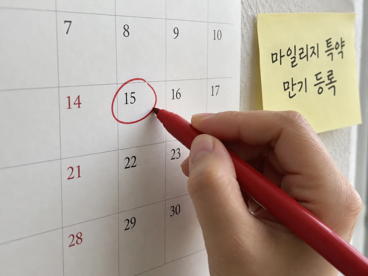 벽걸이 달력 특정 날짜에 빨간 펜으로 동그라미 표시를 하는 손, 옆에 '마일리지 특약 만기 등록'이라고 적힌 포스트잇