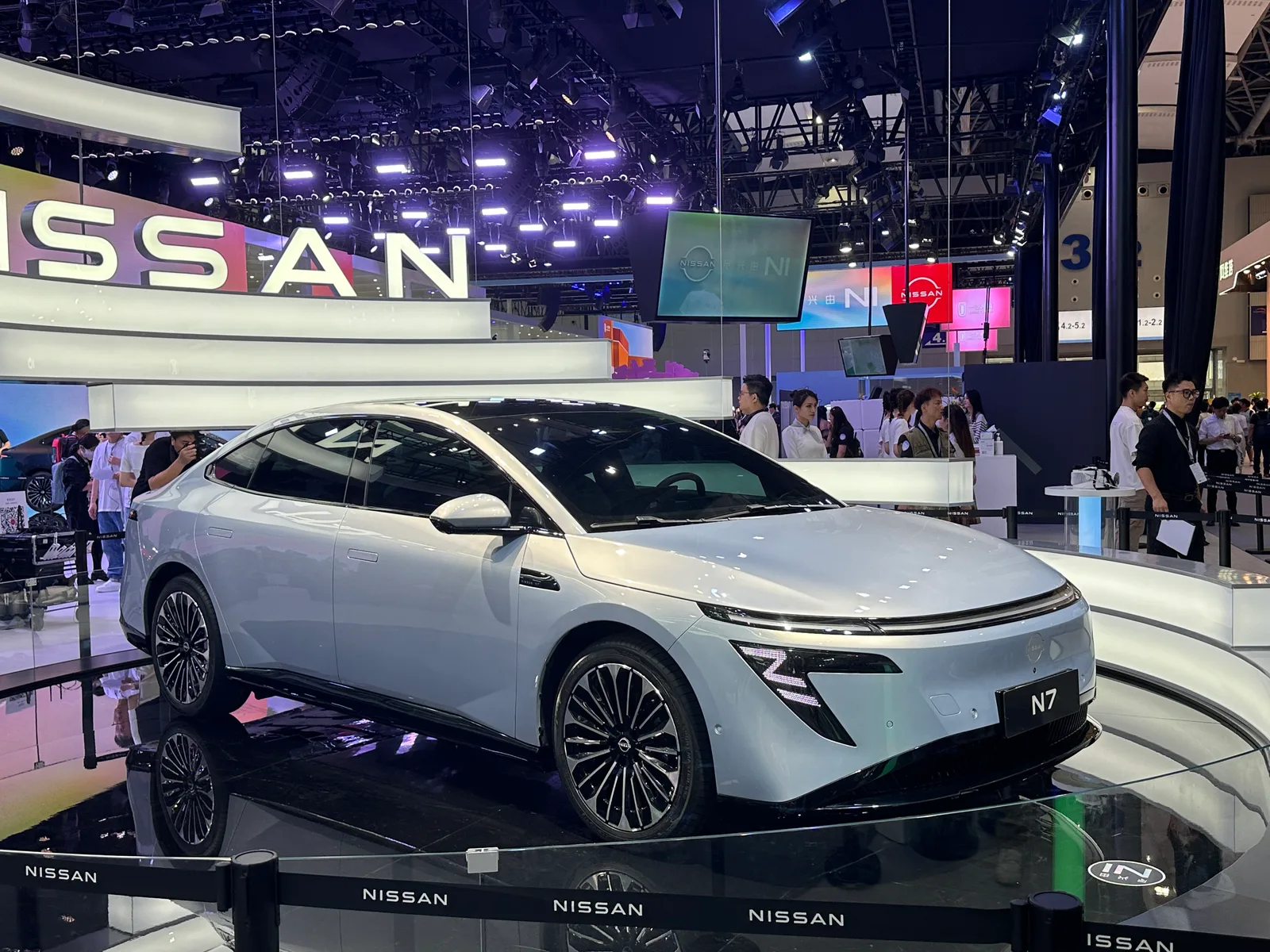 중국 모터쇼 둥펑닛산 부스에 전시된 흰색 닛산 N7 전기 세단의 앞모습, 배경에 대형 NISSAN 로고와 관람객들이 보이는 모습
