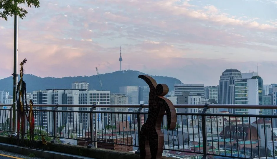 노을 하늘 아래 도심 건물과 멀리 남산 능선이 보이고 난간 앞에 철제 인물 조형물이 서 있는 전망
