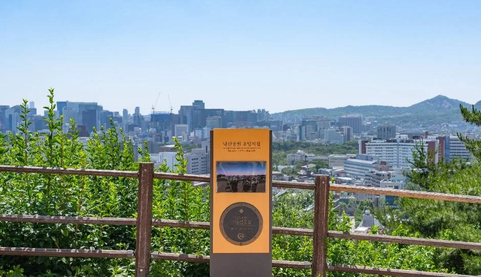 나무 난간 앞에 안내 표지판이 서 있고 멀리 도심 빌딩과 산이 보이는 낙산 전망 지점