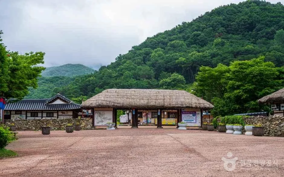 산자락 아래 초가지붕을 얹은 큰 건물과 흙마당이 있는 마을 입구 쪽 풍경